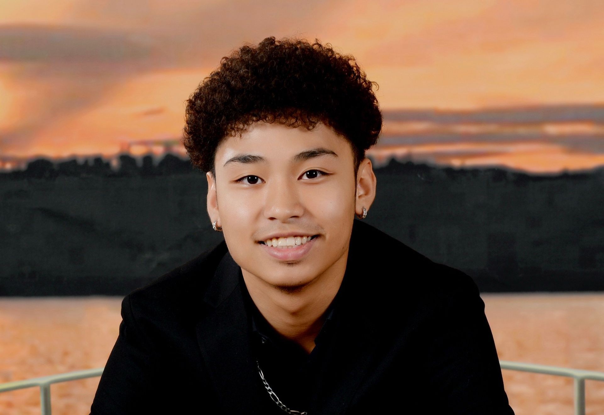 Person smiling at the camera with a sunset backdrop, wearing a black jacket and earrings.